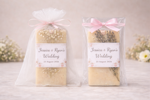 Two wedding soap favor packaging styles side by side: left shows handmade lavender soap in white organza bag with pink ribbon, right shows soap in clear bag with pink satin bow, both featuring custom labels reading 'Wedding 2with event name' with floral design