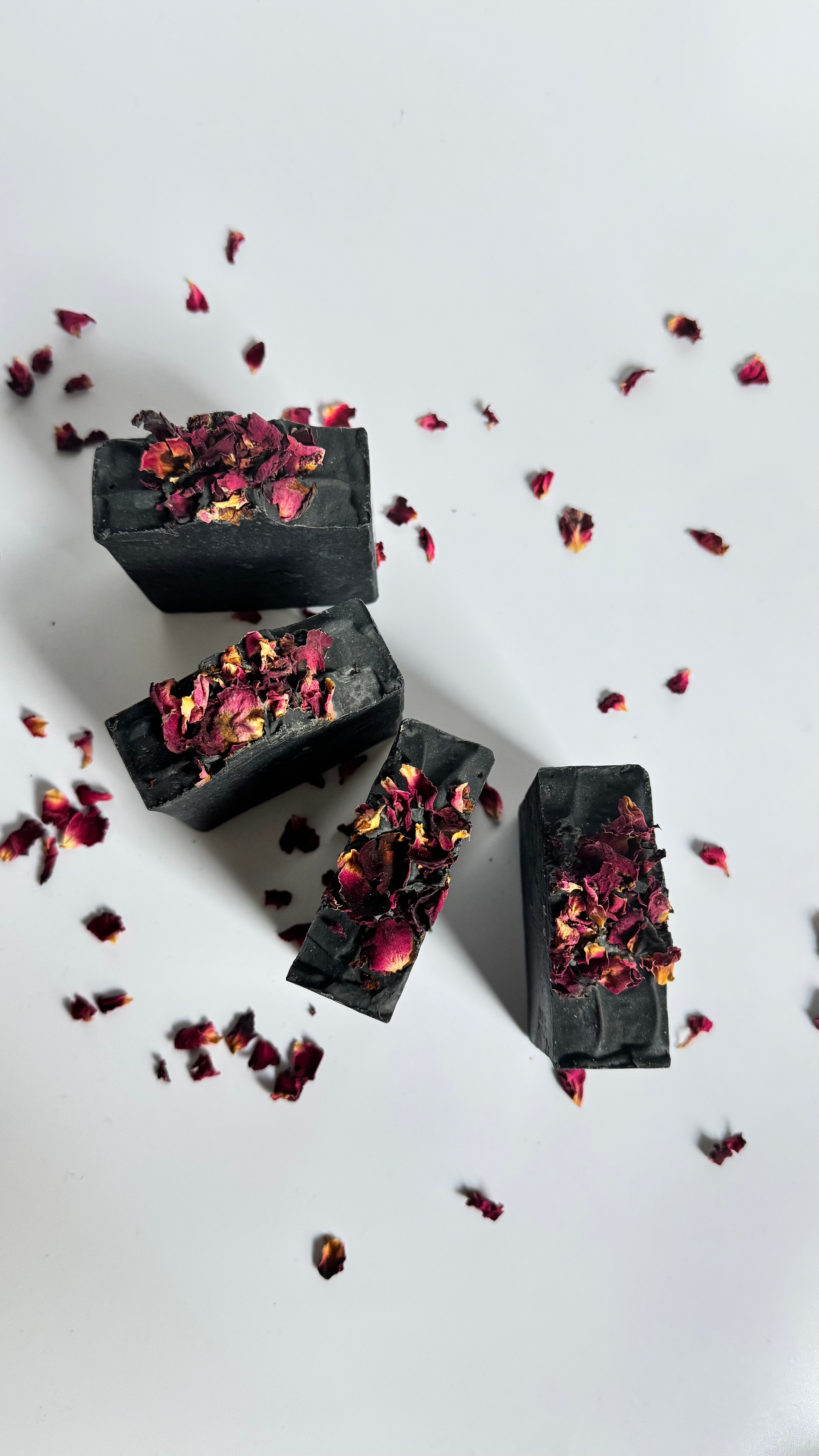 Four black activated charcoal lavender tea tree natural soap bars above view in circle with dried rose petals on a white background