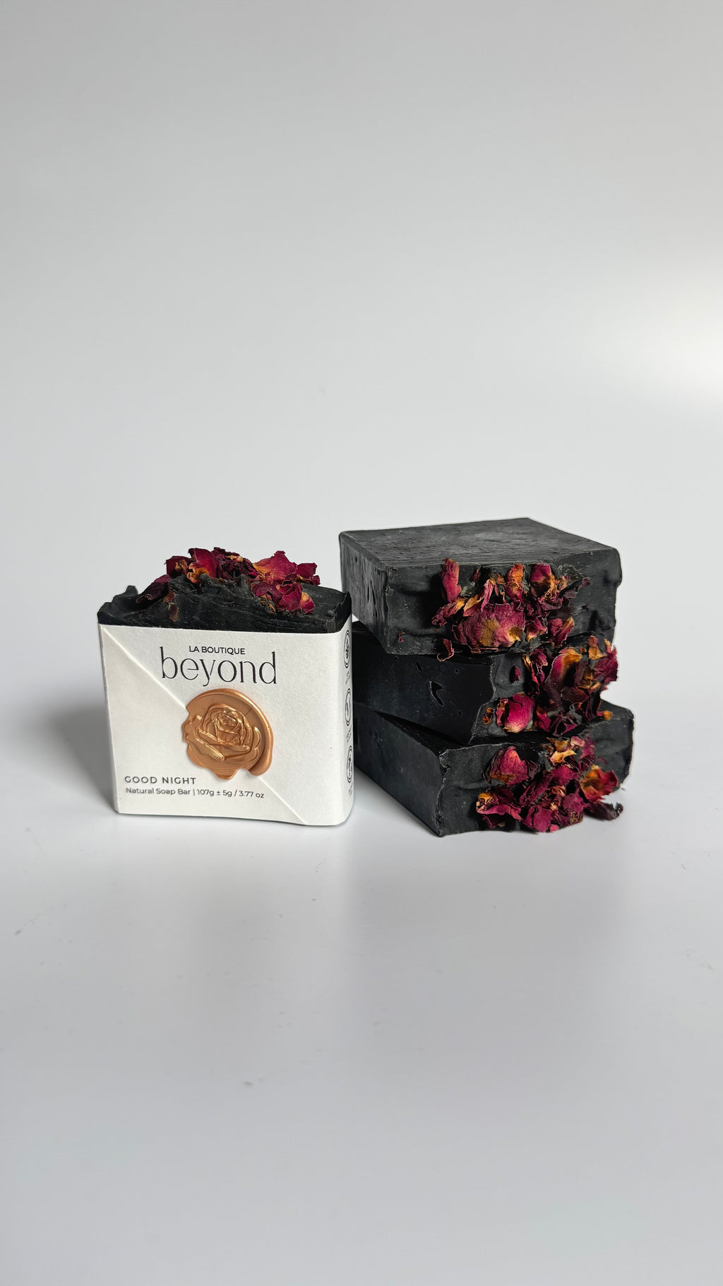 Four black activated charcoal lavender tea tree natural soap bars with dried rose petals and a packaging labeled 'La Boutique Beyond' on a light background.