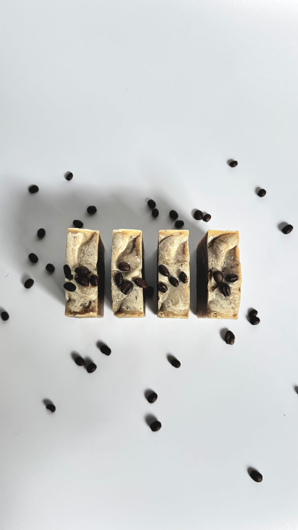 Four brown coffee natural soap bars with coffee beans above view with dried jasmine flowers on a light gray background