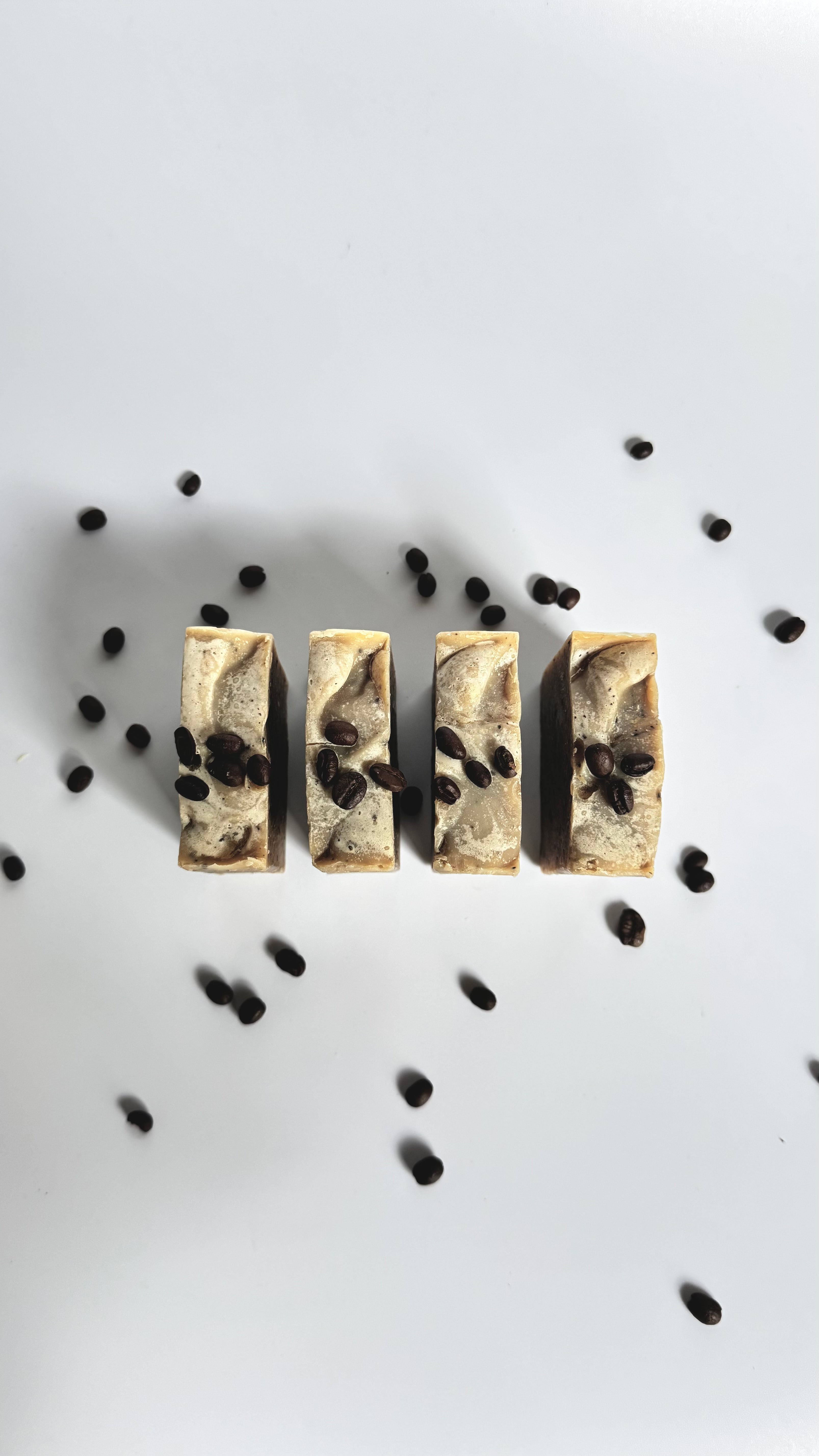 Four brown coffee natural soap bars with coffee beans above view with dried jasmine flowers on a light gray background