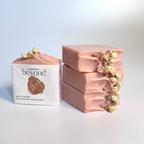 Stack of four pink chamomile rose geranium natural soap bars with dried jasmine flowers and a packaging labeled 'La Boutique Beyond' on a light background.