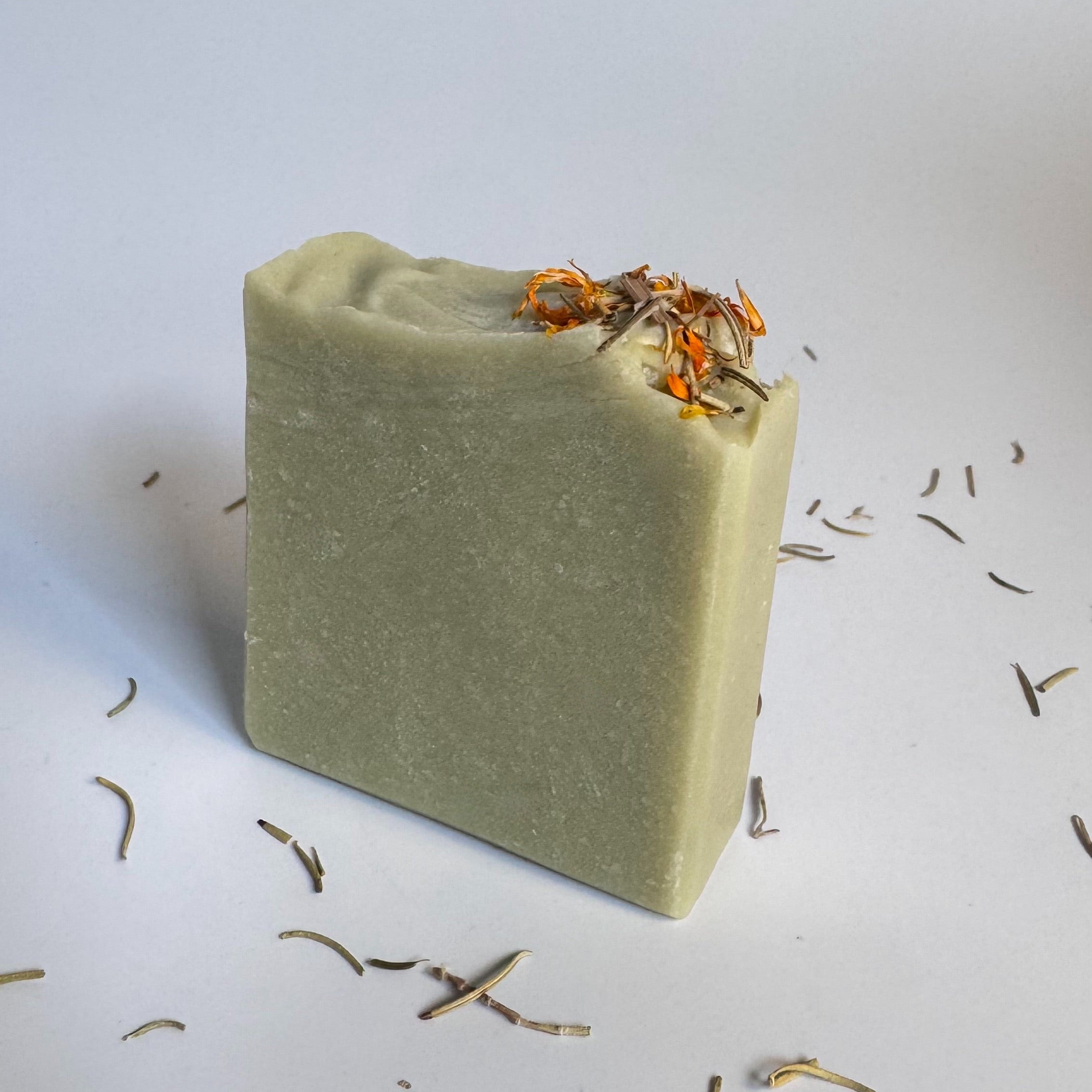 Side view of green rosemary cedarwood natural soap bar with dried floral botanical elements on a light background.