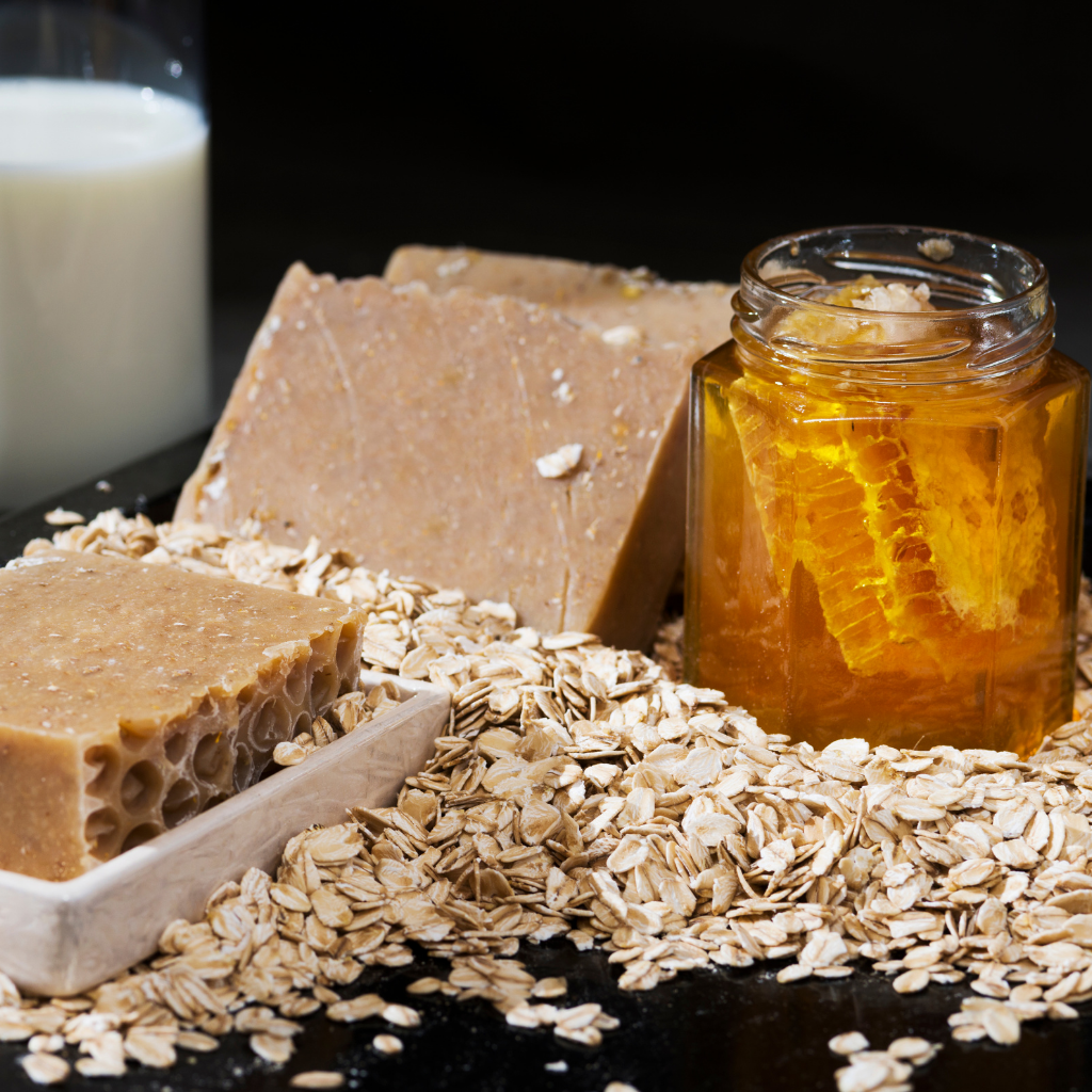 Three pieces of brown soap, a jar of honey, and rolled oats on a dark surface.