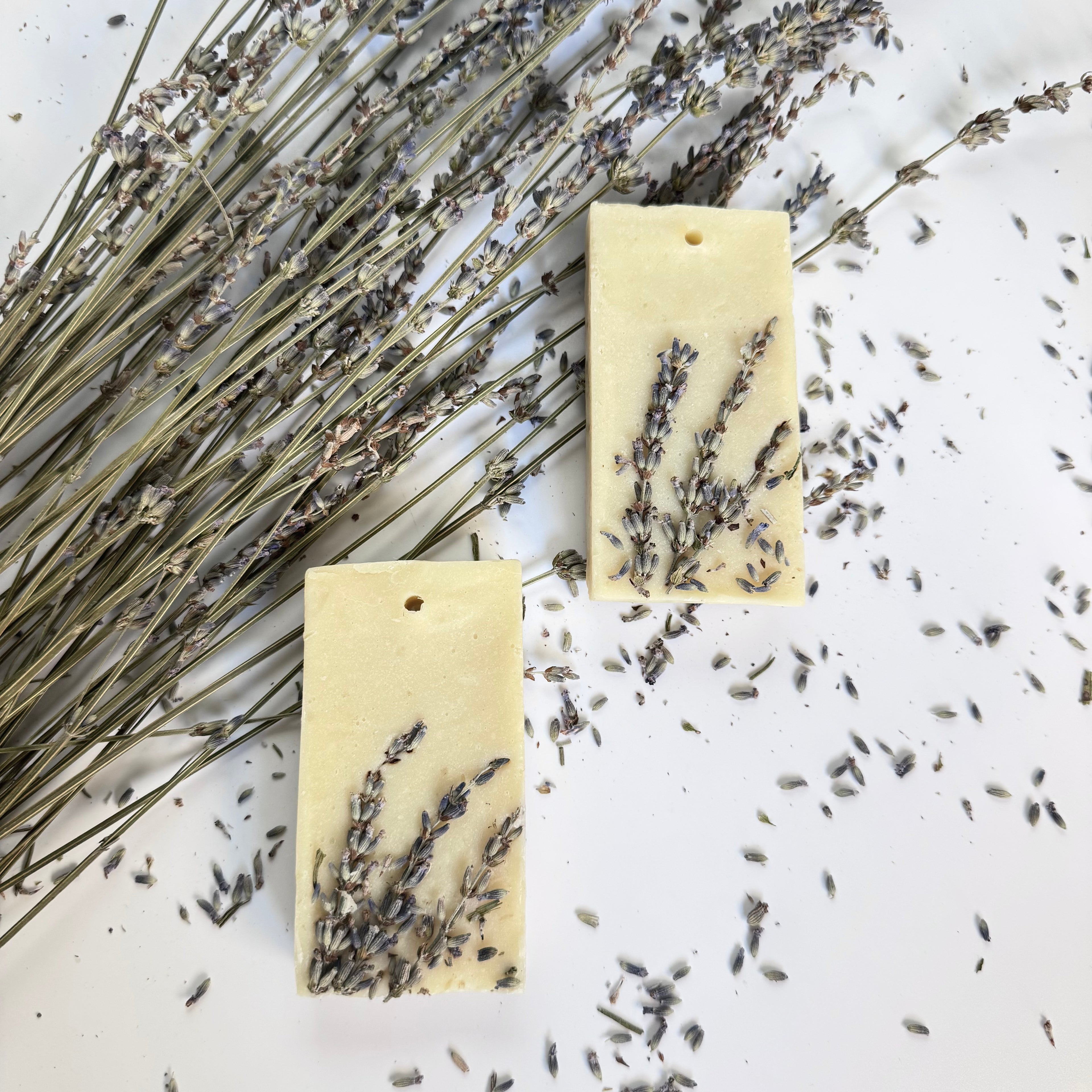 Two rectangular natural hanging soap bars with lavender on a white surface with lavender flowers.