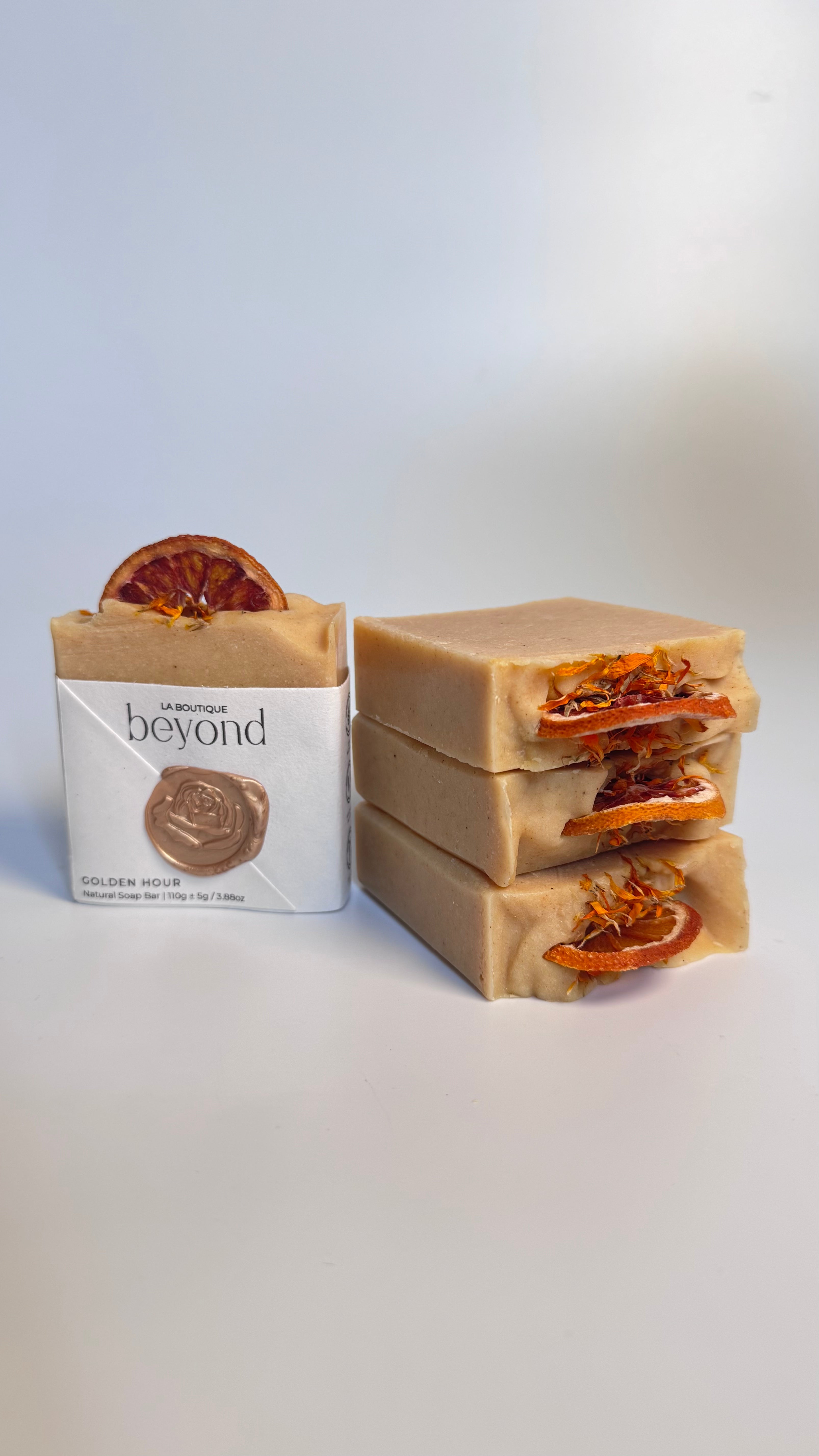 Stack of orange turmeric natural soap bar with dried orange slices on a white background and a packaging labeled 'La Boutique Beyond' on a light background.