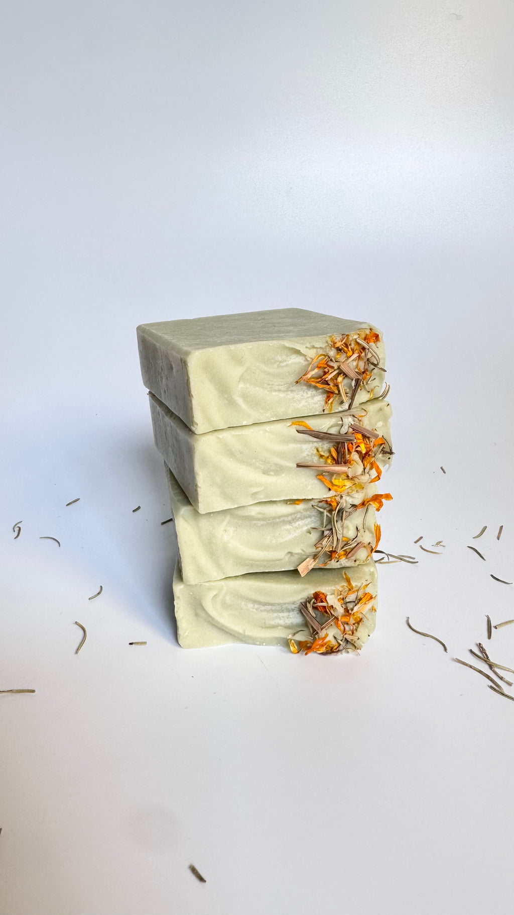 Stack of green rosemary cedarwood natural soap bars with dried floral botanical elements on a light background.