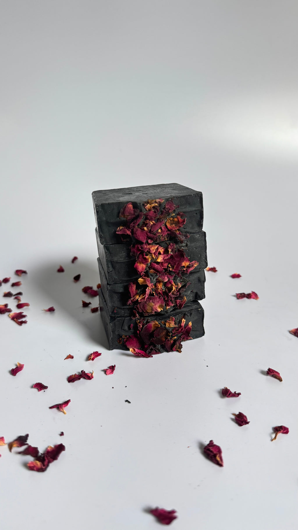 Stack of four black activated charcoal lavender tea tree natural soap bars with dried rose petals on a white background 