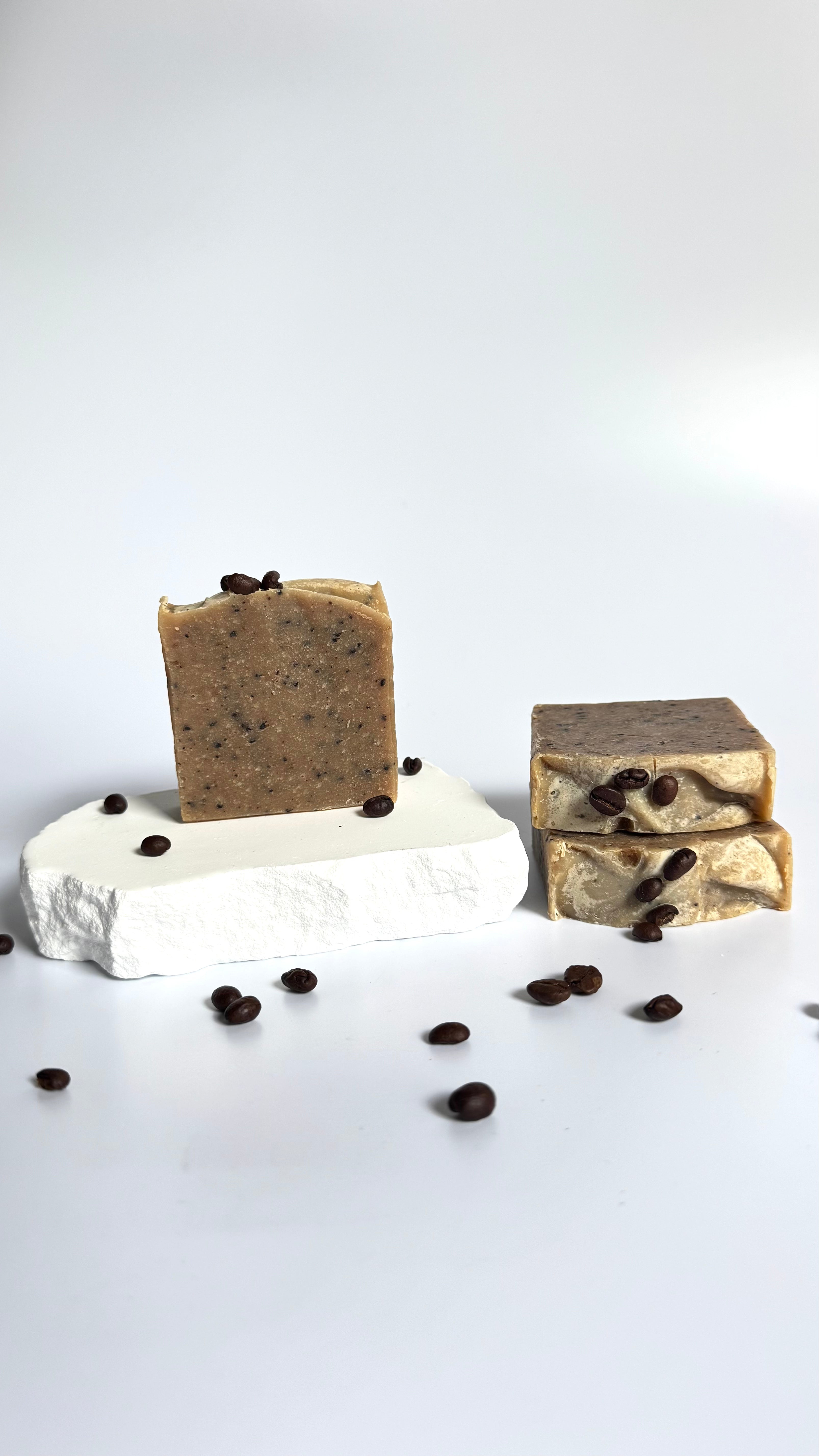 Stack of three brown coffee natural soap bars with coffee beans with dried jasmine flowers on a light gray background