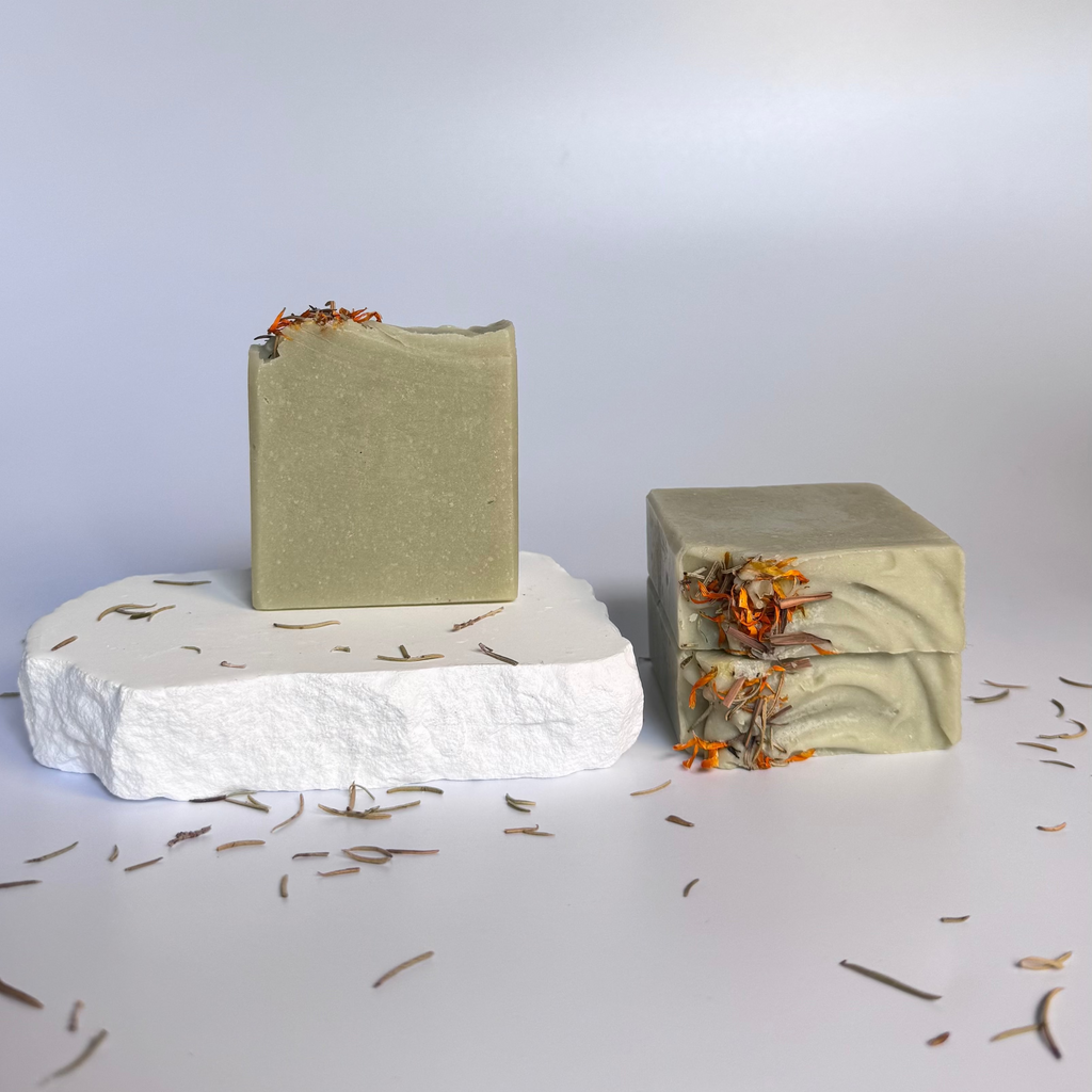 Three green rosemary cedarwood natural soap bars with dried floral botanical elements on a light background.