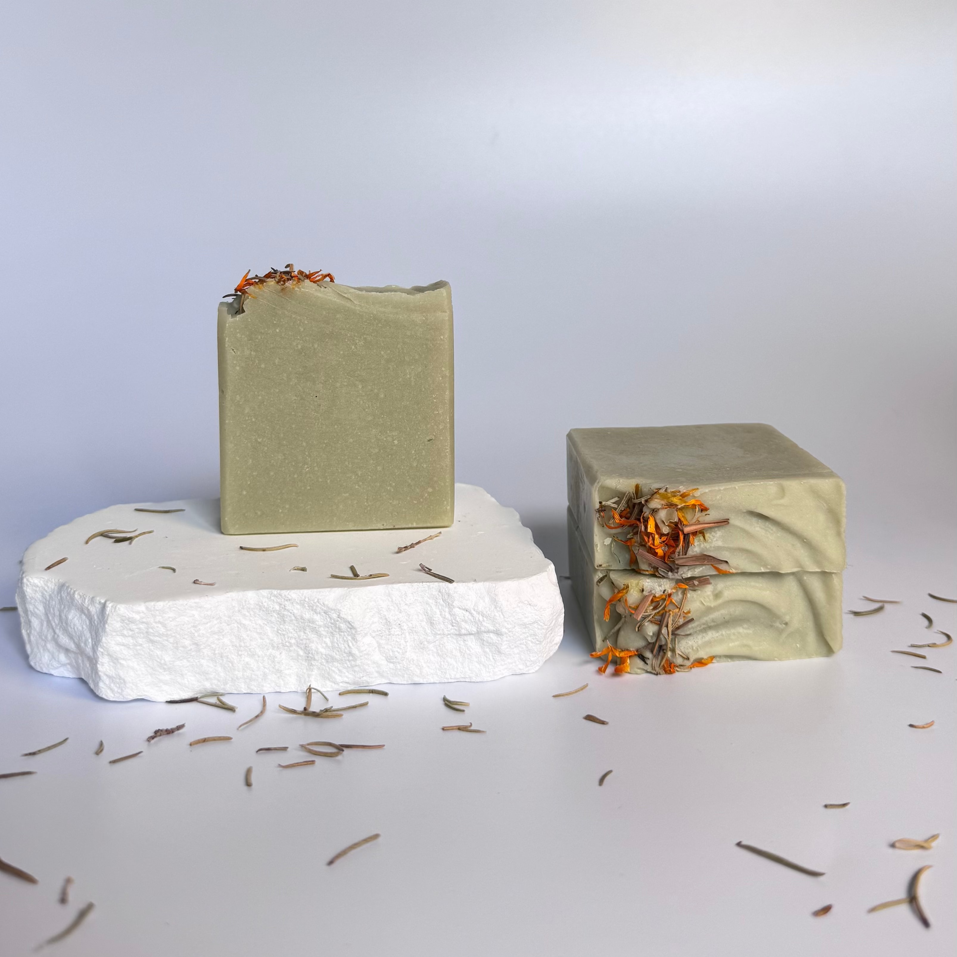 Three green rosemary cedarwood natural soap bars with dried floral botanical elements on a light background.