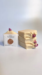 White beige cream coconut milk natural soap bars with dried pink roses on a white surface and a packaging box labeled 'La Boutique Beyond' on a light background.