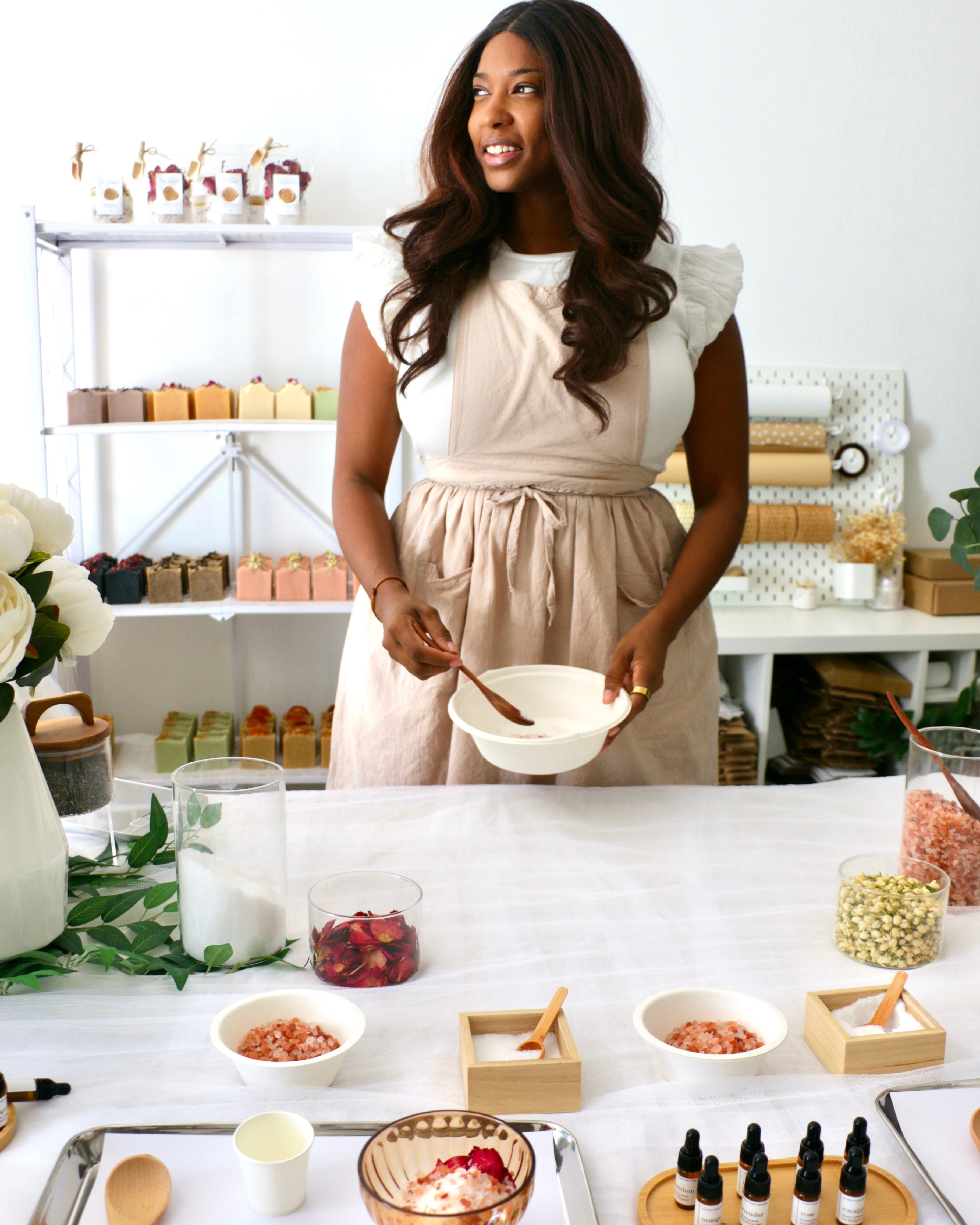 Founder Cathy at La Boutique Beyond at a table setup for live bath soak station in Dubai with various bowls, natural ingredients, and essential oils, and utensils on a white surface.