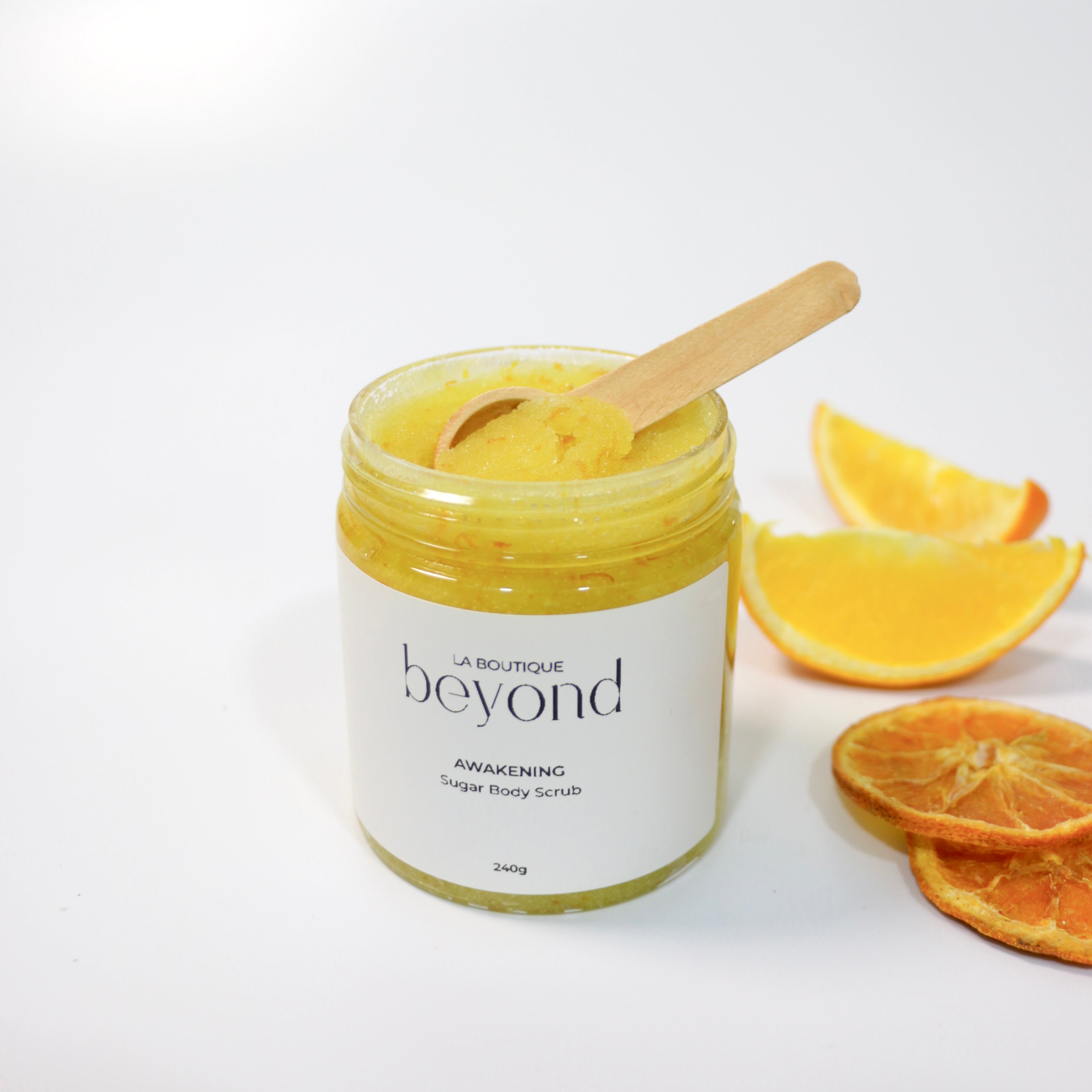 Jar of 'La Boutique Beyond Awakening' orange sugar body scrub with a wooden spoon and orange slices on a white background