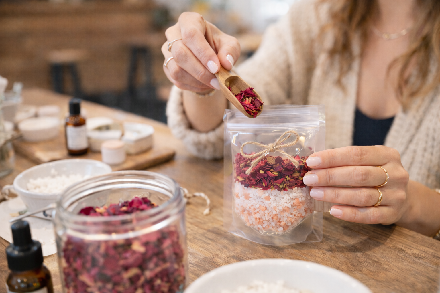 Person holding a container of bath soak made with dried herbs, essential oils with a wooden spoon on a wooden table.